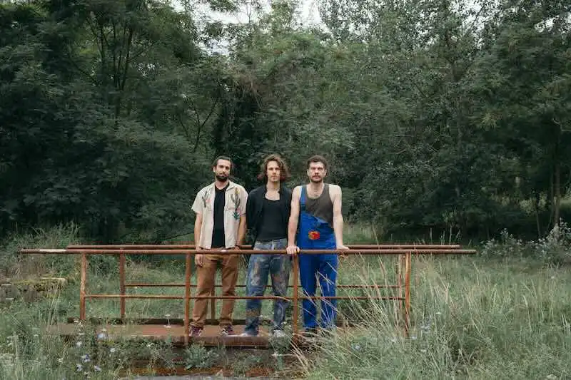 Three men standing on a small bridge in nature, representing diversity and unity in music scenes.