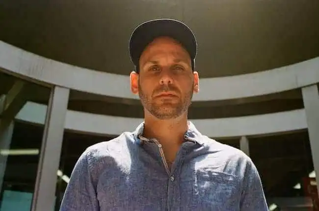 Male model wearing a cap and casual shirt outdoors in bright sunlight.