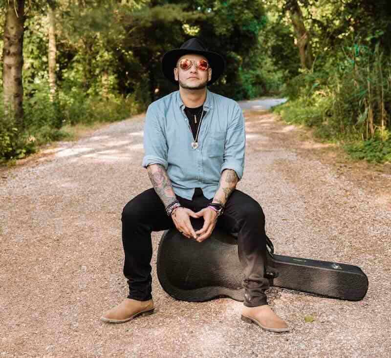 Individual with guitar case in a forested setting.