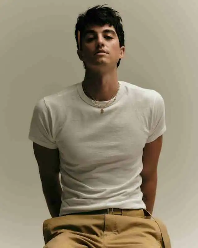 A confident young man with dark hair in a white t-shirt and beige pants, showcasing casual, modern fashion.