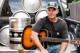 A man with a guitar sitting on the tailgate of a truck near beer kegs and a pint, outdoors, casual vibe.