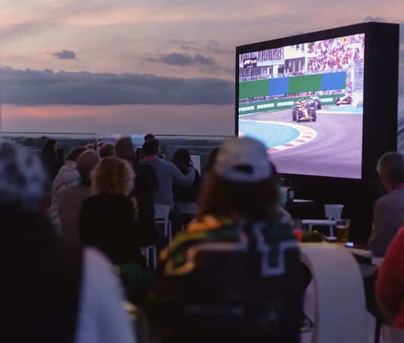 Outdoor motorsport fans watch Formula 1 on large screen.
