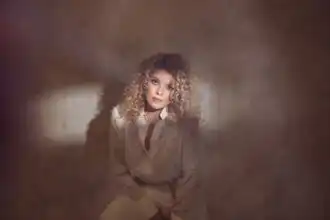 Portrait of a woman with curly blonde hair, wearing a beige shirt against a soft, warm background.