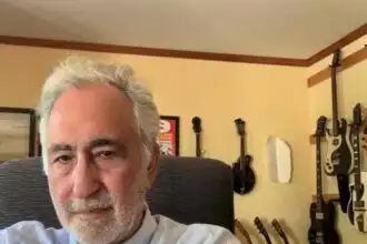A mature man with white hair and beard poses in a room with multiple guitars on the wall, showcasing a love for music and literature.