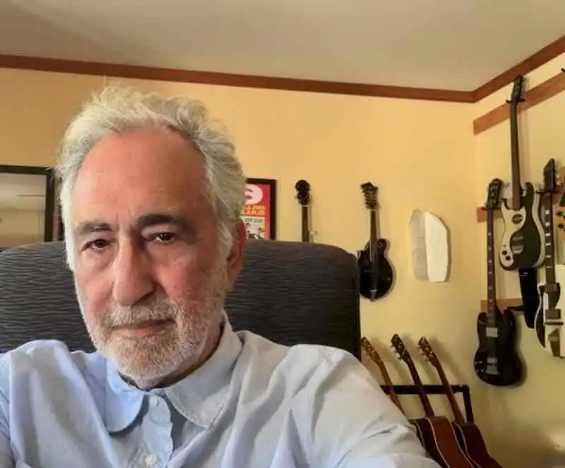 A mature man with white hair and beard poses in a room with multiple guitars on the wall, showcasing a love for music and literature.