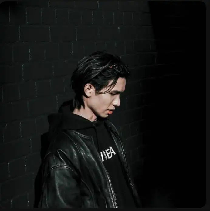 Young man deep in thought, standing by a dark brick wall, wearing a leather jacket and hoodie.