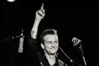 Vintage male musician on stage with guitar, celebrating with raised finger, black-and-white image.
