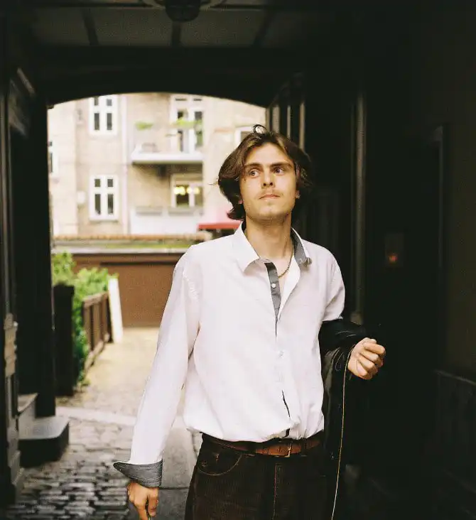A young man with wavy brown hair wearing a white shirt and holding a jacket, standing in an outdoor passageway with an urban background.