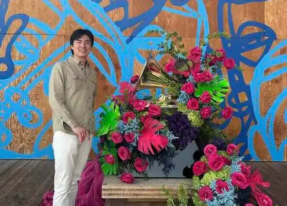 Smiling man standing next to colorful flower arrangement on grand piano at an art exhibit.