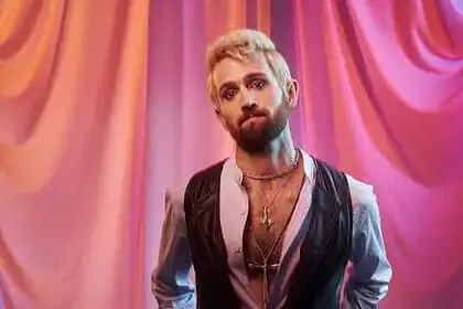 Male artist with a beard and styled hair standing in front of colorful draped curtains.