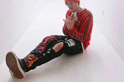 Young man sitting on the floor in a white hallway, wearing a red beanie and colorful streetwear.
