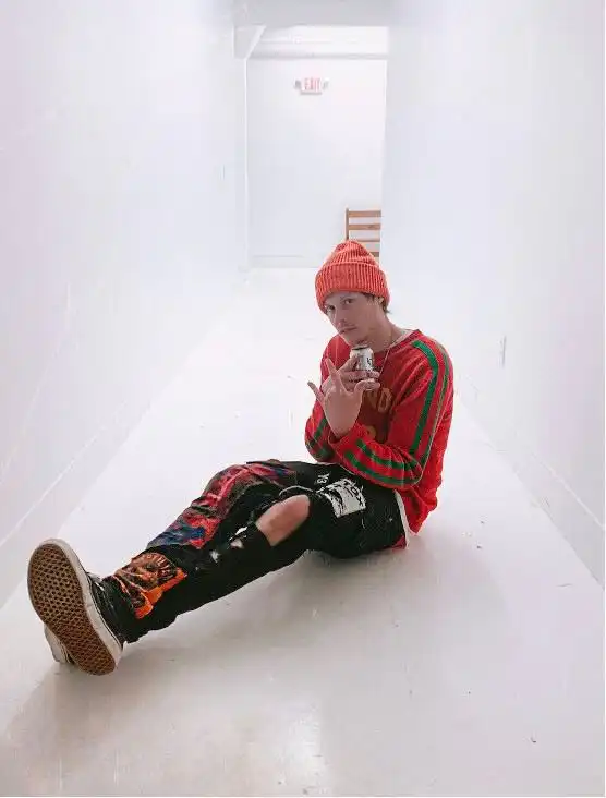 Young man sitting on the floor in a white hallway, wearing a red beanie and colorful streetwear.