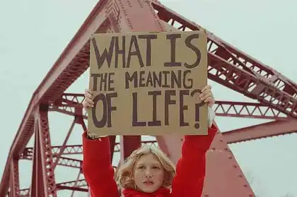 Woman holding a sign asking "What is the meaning of life?" under a bridge.