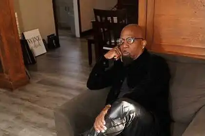 Alxandr sitting thoughtfully on a sofa, wearing stylish black leather pants and boots, in a cozy ind.