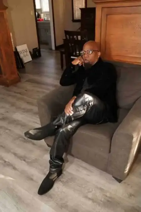 The Alxandr – Tender Love (New Single) Alxandr sitting thoughtfully on a sofa, wearing stylish black leather pants and boots, in a cozy ind.