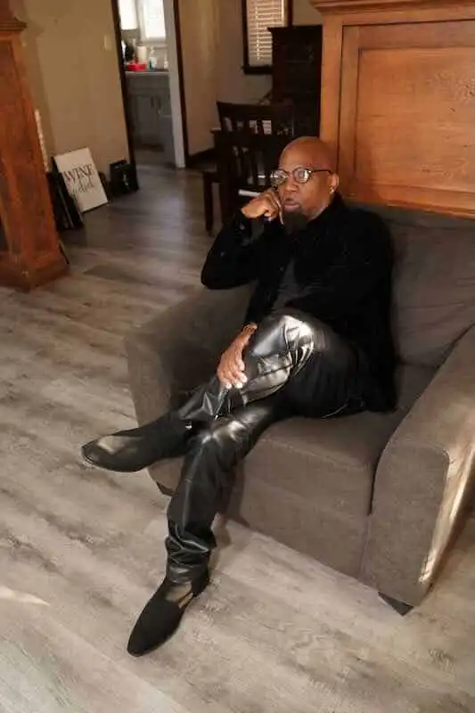 The Alxandr – Tender Love (New Single) Alxandr sitting thoughtfully on a sofa, wearing stylish black leather pants and boots, in a cozy ind.