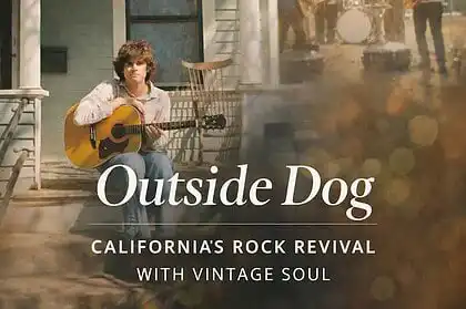 Young man playing acoustic guitar on porch during outdoor music event in California.