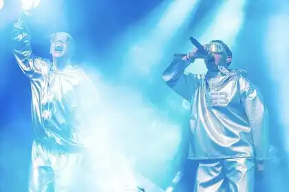 VÆB and GamerBoi performing on stage with futuristic outfits and microphones.
