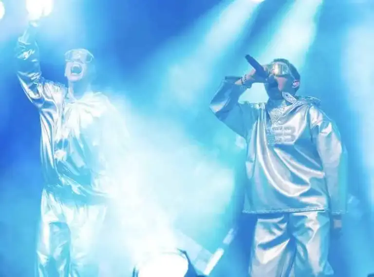 VÆB and GamerBoi performing on stage with futuristic outfits and microphones.
