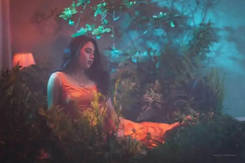 A woman in an orange dress sits peacefully among vibrant greenery and soft lighting in a tranquil garden scene.