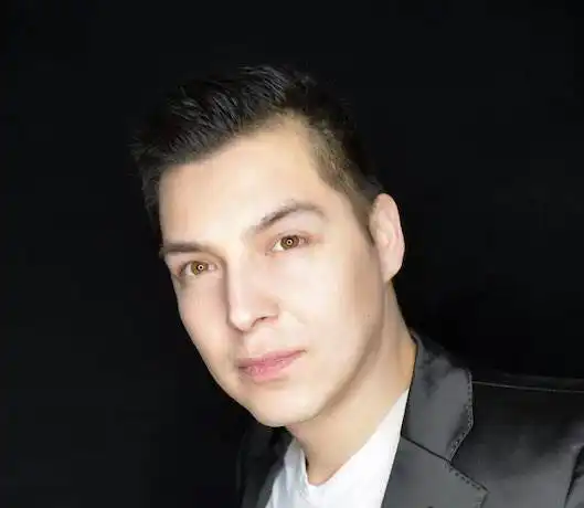 Close-up of a young man with dark hair and fair skin against a black background, wearing a black blazer.