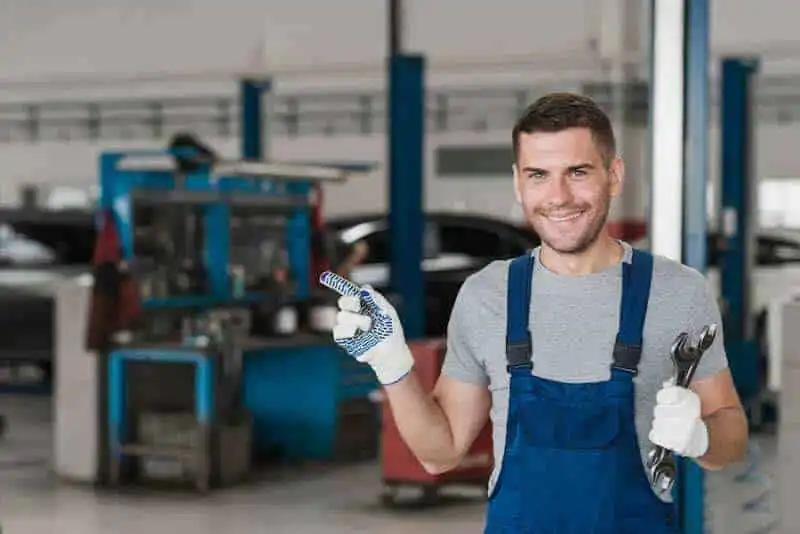 A smiling male mechanic holding a wrench and a spark plug in an auto repair shop.