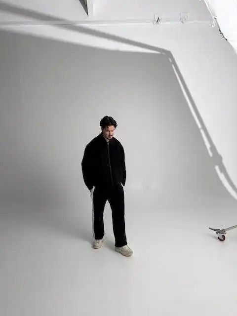 A young man dressed in casual black attire standing on a white studio floor with a skateboard nearby.