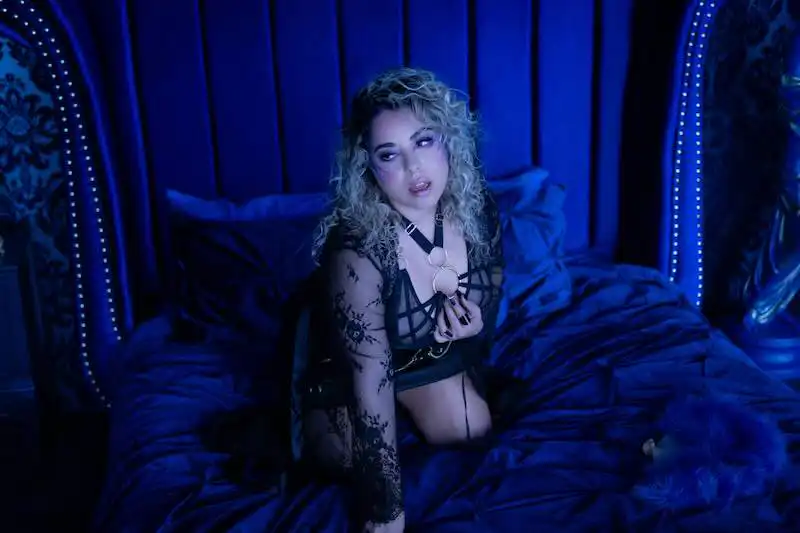 A woman with curly hair in black lace lingerie, sitting on a luxurious blue bed in a dimly lit bedroom.