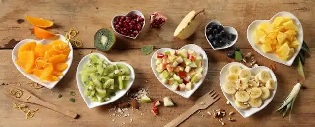 Colorful fresh fruit salad with bananas, kiwis, melons, berries, and apples in heart-shaped bowls.