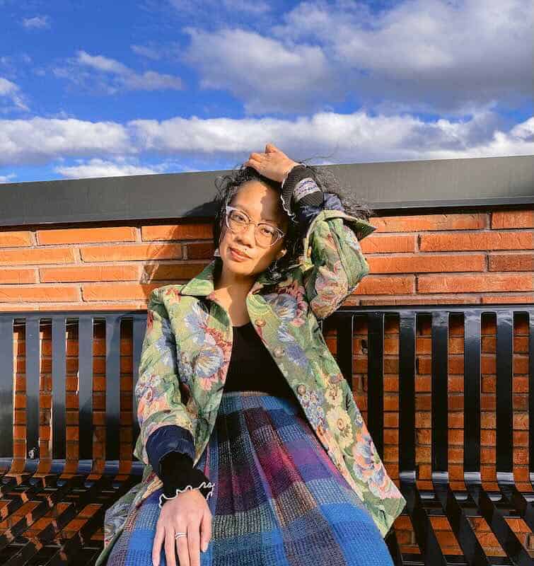 A young woman with glasses posing outdoors on a bench, wearing a colorful patchwork skirt and floral jacket under a bright sky.