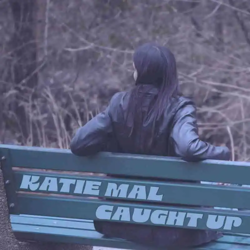 A woman with dark hair in a leather jacket sits on a park bench, looking away into the distance, surrounded by nature.
