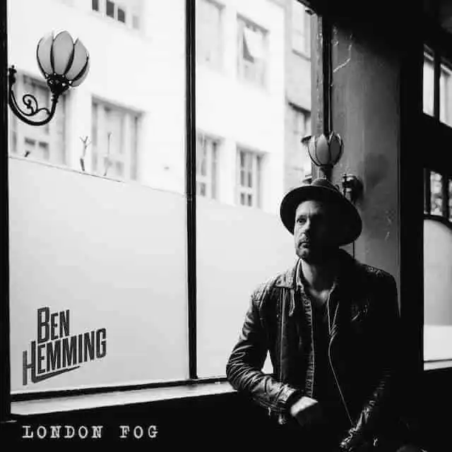 A black and white photo of Ben Hemming in London fog, wearing a hat and leather jacket, sitting by a window.