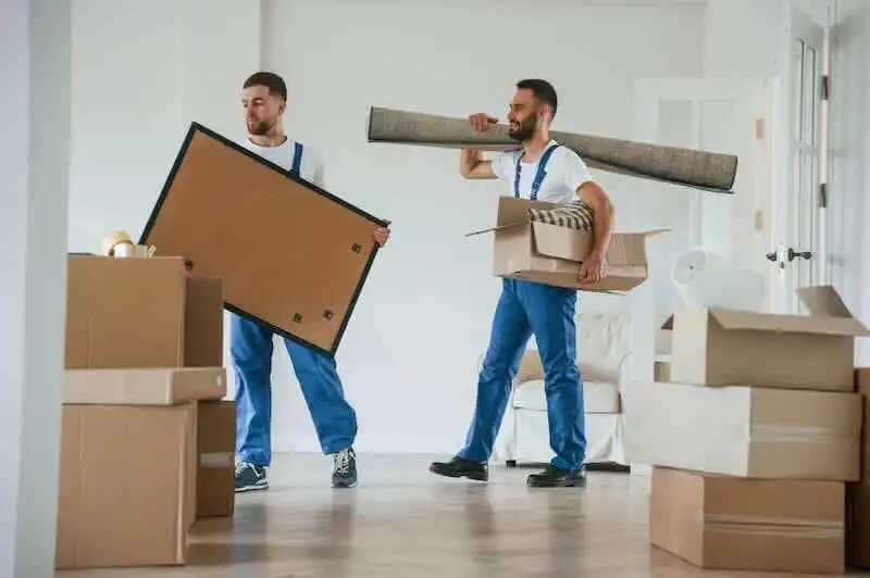 Two men packing belongings into boxes for a move, carrying large items like a rolled carpet and picture frame in a bright, modern home.