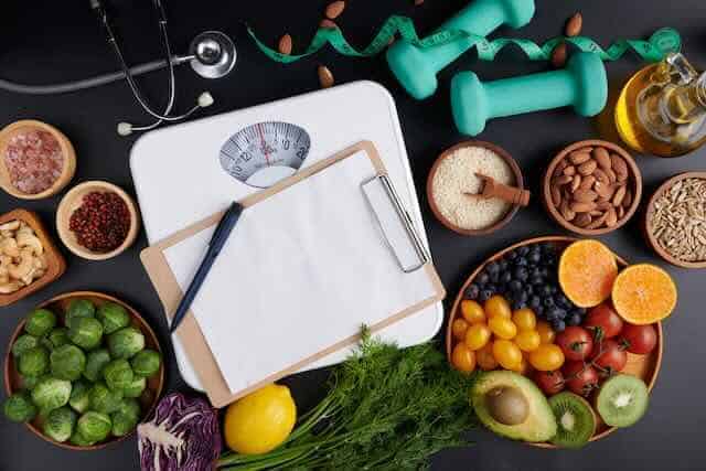 A variety of fresh fruits, vegetables, and nuts displayed alongside scales and dumbbells for balanced dieting and fitness.