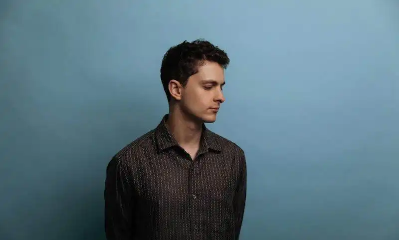 Young man with dark hair in a patterned shirt posing against a blue background, reflecting introspection.